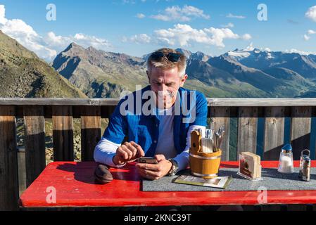 Ein Wanderer sitzt auf der Terrasse der Berghütte Brunnenkogelhaus und schaut auf sein Handy vor den Ötztaler Alpen und dem Gurgltal in Österreich Stockfoto