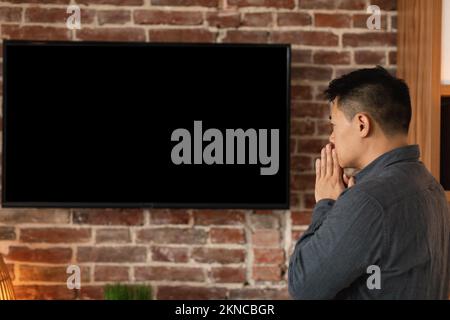 Trauriger besorgter asiatischer Mann mittleren Alters, der Nachrichten im großen fernseher mit leerem Bildschirm im Innenbereich des Heimbüros sieht, Profil Stockfoto