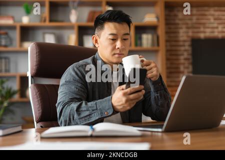 Ein sehr beschäftigter, reifer chinesischer Geschäftsmann mit Computer liest Nachrichten auf dem Smartphone und trinkt Kaffee Stockfoto