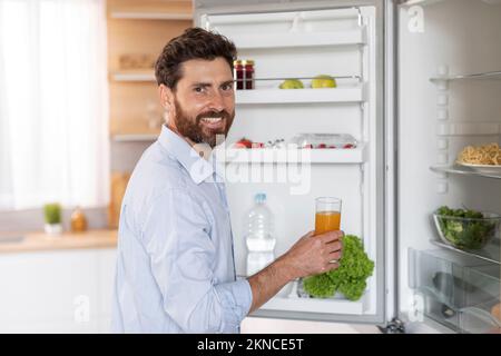 Ein glücklicher, gutaussehender, weißer Mann mit Bart im Hemd öffnet die Kühlschranktür und nimmt ein Glas Saft Stockfoto