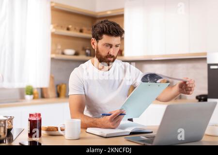 Ein erwachsener, weißer Mann mit Bart arbeitet mit Dokumenten und Laptop in einer minimalistischen Küche Stockfoto