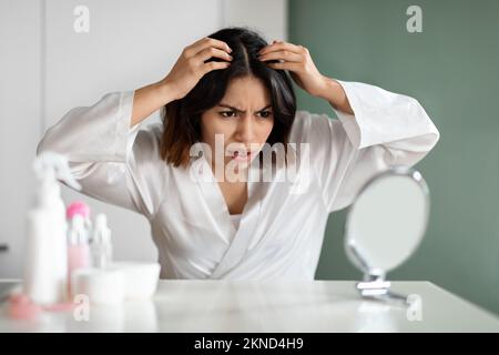 Wütende Frau im weißen Bademantel, die im Schlafzimmer sitzt und Haare überprüft Stockfoto