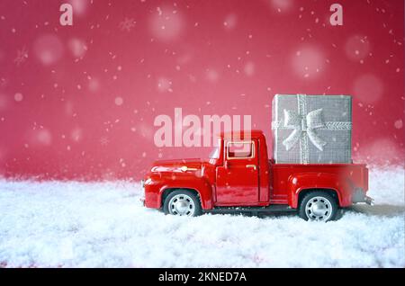 Weihnachtlich-roter Hintergrund mit Schnee und Retro-rotes Auto mit Geschenkbox. Frohe Weihnachten und frohe Neujahrsgrüße Winterkarte mit Kopierbereich. Feiertage Stockfoto