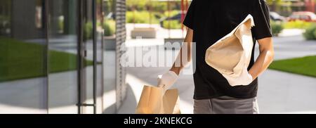 Kontaktlose Lieferung während des COVID-19 Pandemiesperrkonzepts. Der Kurier trägt eine Maske und Handschuhe und hält einen Paketkasten und prüft die Adressdetails auf dem Stockfoto