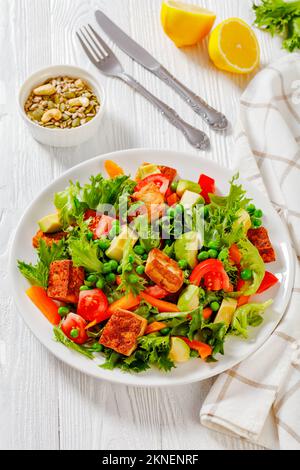 Tofu-Salat mit Avocado, knusprigen Salatblättern, grünen Erbsen, Tomaten, gelbem süßen Pfeffer auf weißer platel, vertikaler Blick von oben Stockfoto