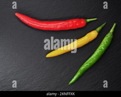 Drei verschiedenfarbige Chili-Paprika auf schwarzem Schieferhintergrund, Draufsicht auf rote, gelbe und grüne Chilischoten Stockfoto