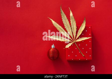 Festlicher roter weihnachtshintergrund mit goldenem Marihuana-Blatt und Geschenkbox. Weihnachtsdekor und goldenes Marihuana, rotes Weihnachtskonzept für poste Stockfoto