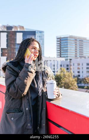 Attraktive hispanische Frau mit Thermoskanne und heißem Kaffee, die telefoniert, während sie in der Nähe der Stadtgrenze mit Gebäuden steht Stockfoto