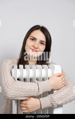 Eine braune Frau umarmt einen elektrischen Ölwärmer und wärmt sich im Winter auf. Stockfoto