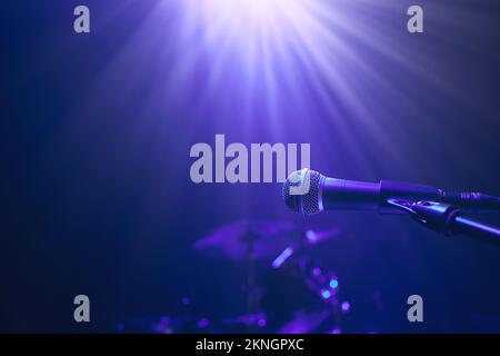 Selektiver Fokus auf beleuchtetes Mikrofon gegen Trommelkit auf der Bühne in blauem Licht. Stockfoto