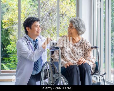 Lächelnd positiver asiatischer Mann Arzt im weißen Mantel spricht mit Sorgfalt zu älteren älteren älteren weiblichen Patienten, die in einem Rollstuhl in der medizinischen Praxis in der Zusammenarbeit sitzen Stockfoto