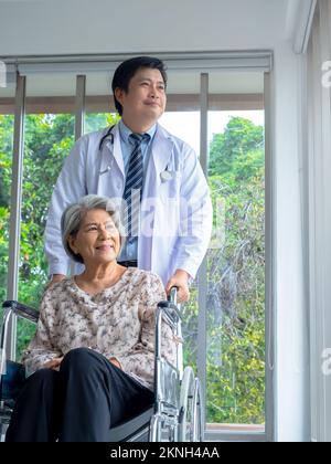 Lächelnd glückliche asiatische ältere ältere Patientin sitzt im Rollstuhl mit Mann Arzt in weißem Mantel steht in der Arztpraxis im Krankenhaus, vertikale St. Stockfoto