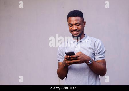 Ein fröhlicher afroamerikanischer Mann, der sein Handy auf grauem Hintergrund benutzt Stockfoto