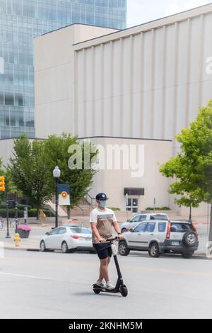 07 25 2020 Tulsa USA - die neue Normalität - Ein junger Mann in Shorts, T-Shirt, Ballkappe und Gesichtsmaske fährt mit einem elektrischen Roller die Straße hinunter Stockfoto