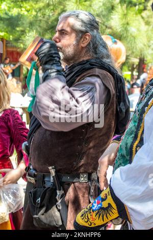 Ein Mann in Kostümen mit langen grauen Haaren trinkt zusammen mit anderen Fair-Stars im Hintergrund beim Oklahoma Renaissance Festival Muskogee Okla einen Drink aus einem Holzbecher Stockfoto