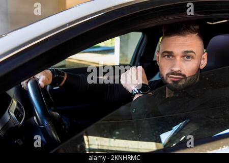 Ein weißer Mann im Anzug, mit einer Uhr und einem Armband hinter dem Steuer des Autos. Schaut aus dem Fenster. Stockfoto