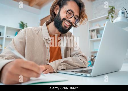 Fröhlicher junger kreativer Unternehmer, der lächelnd lächelt, schreibt in seinem Büro neben dem Laptop ein Notizbuch Stockfoto