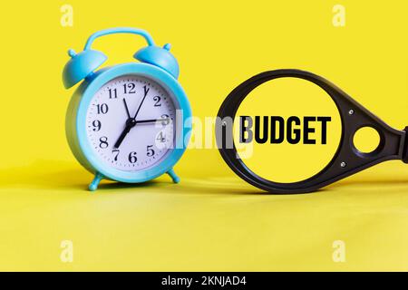 DAS Wort BUDGET auf einer Lupe auf gelbem Hintergrund mit Uhr Stockfoto