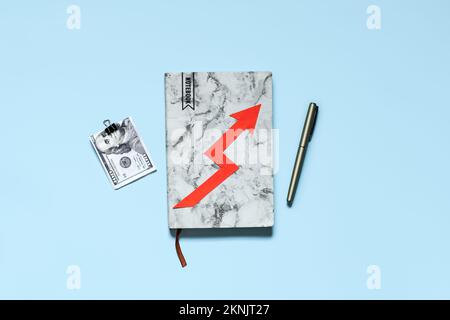 Steigende Lebenshaltungskosten, Inflation, Finanz- und Wirtschaftskrise. Notizbuch mit Geldscheinen und rotem Pfeil auf blauem Hintergrund. Stockfoto