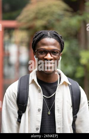 Fröhlicher afrikanischer Brillenschüler, der vor der Kamera lächelt, steht in der Nähe des Universitätscampus draußen Stockfoto