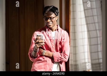 Ein optimistischer afroamerikanischer Mann steht neben dem Fenster im Zimmer und lächelt am Telefon Stockfoto