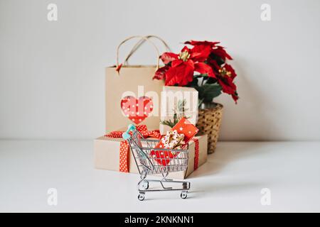 Weihnachtseinkäufe und Weihnachtsverkauf. Weihnachtlicher Mini-Einkaufswagen mit roter Geschenkbox, Poinsettias Xmas Blume und Craft-Einkaufstasche auf Weiß Stockfoto