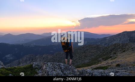 Eine Wanderin steht auf einem großen Felsen mit den Bergen im Hintergrund und beobachtet den Sonnenuntergang. Stockfoto