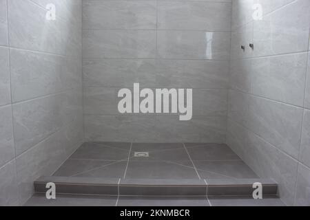 Moderne Dusche nach Renovierung. Sideboard, Abfluss im Boden und Zusatz für Wasserhähne in der Wand. Graue Fliesen auf dem Boden und an den Wänden Stockfoto