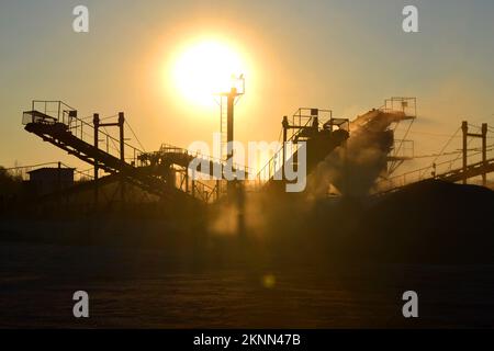 Bergbau. Zerstäuber im Granitbruch bei Sonnenaufgang. Brecher im Steinbruch. Viel Staub. Steinschlag. Industrielle Kies. Kies-Förderer. Kegelsteinbrecher, strahlende Sonne Stockfoto