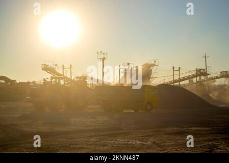 Bergbau. Zerstäuber im Granitbruch bei Sonnenaufgang. Brecher im Steinbruch. Viel Staub. Steinschlag. Industrielle Kies. Kies-Förderer. Kegelsteinbrecher, strahlende Sonne Stockfoto