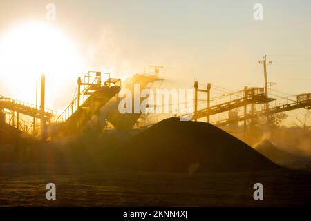 Bergbau. Zerstäuber im Granitbruch bei Sonnenaufgang. Brecher im Steinbruch. Viel Staub. Steinschlag. Industrielle Kies. Kies-Förderer. Kegelsteinbrecher, strahlende Sonne Stockfoto