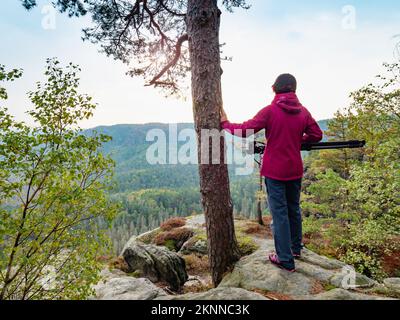Die Fotografin in roter Jacke bleibt mit der Kamera auf dem Stativ auf der Klippe und denkt nach. Eine Frau hält den Baum am Rand. Traumhafte neblige Landschaft, neblige Su Stockfoto