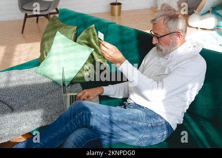 Älterer Mann, der Online-Zahlungen mit einem Laptop abgibt. Alter Großvater, der E-Commerce mit Kreditkarte kauft. Rentner mit Debitkarte für Stockfoto