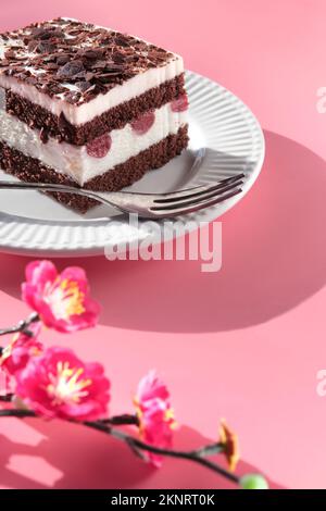 Schokoladenkuchen mit Sauerkirschen. Ein Stück Kuchen auf einem Teller mit Gabel. Süßes Dessert auf pinkfarbenem Hintergrund mit leuchtenden Pflaumenblüten in Magenta. Stockfoto