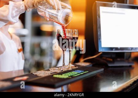 Der Barkeeper gießt Rotwein aus einem Messbecher an der Theke in zwei langstielige Weingläser. Unscharfer Hintergrund. Party, Cocktail, Alkohol Stockfoto