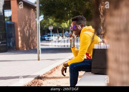 Ein junger Afrikaner in einem gelben Sweatshirt, Jeans und Sonnenbrille, der auf einer Straßenbank sitzt und mit seinem Handy neben einer Einkaufstasche spricht Stockfoto
