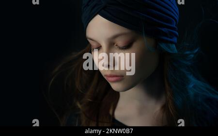 Hübsches kleines Teenager-Mädchen mit meditativem Gesichtsausdruck in mehrfarbigen Lichtern. Das dunkle Porträt eines Kindes mit Turban und fließendem Haar Stockfoto