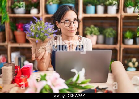 Junge hispanische Frau, die im Blumenladen arbeitet und Videoanrufe macht, konzentrierte sich auf Zweifel mit dem Finger am Kinn und schaute auf und fragte sich Stockfoto