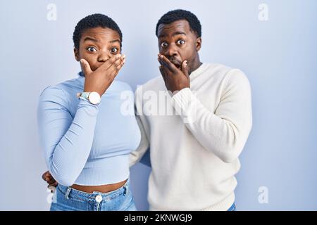 Junges afroamerikanisches Paar, das über blauem Hintergrund steht, schockiert, den Mund mit Händen für einen Fehler zu verschließen. Geheimes Konzept. Stockfoto