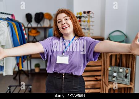 Junge rothaarige Frau, die als Managerin in einer Boutique arbeitet und in die Kamera schaut und mit offenen Armen zum Umarmen lächelt. Fröhlicher Ausdruck, der sich über die Zukunft hinwegsetzt Stockfoto