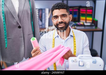 Ein junger arabischer Schneider, der selbstbewusst Stoff und Faden in der Schneiderei hält Stockfoto