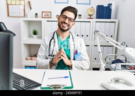 Junger Mann mit Bart trägt Arztuniform und Stethoskop in der Klinik Hände zusammen und Daumen gekreuzt lächelnd entspannt und fröhlich. Erfolg und Stockfoto