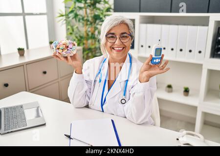 Eine Frau mittleren Alters mit grauen Haaren, die eine Arztuniform trägt und das Blutzuckermessgerät hält und mit einem fröhlichen und kühlen Lächeln im Gesicht lächelt. Zähne zeigen. Stockfoto