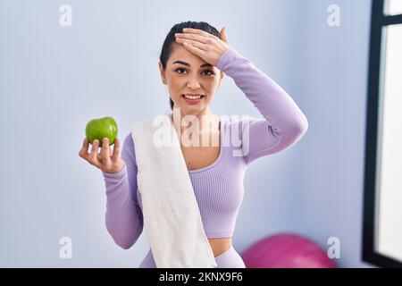 Junge Frau in Sportkleidung und Handtuch, die grünen Apfel isst, gestresst und frustriert von Hand auf Kopf, überrascht und wütend im Gesicht Stockfoto