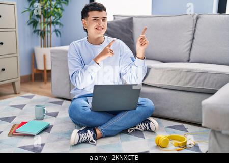 Nicht binäre Person, die mit einem Computer-Laptop studiert und lächelt und mit beiden Händen und Fingern auf die Kamera zeigt Stockfoto