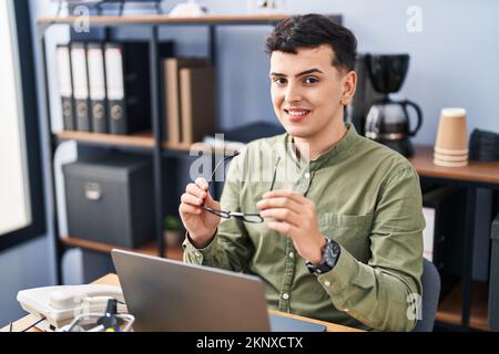 Junger nicht-binärer Mann, der im Büro eine Brille mit einem Laptop hält Stockfoto