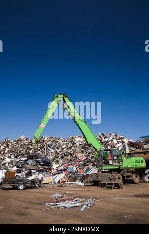 Kranlader mit Greiferarm und Magnet-Entladeanhänger vor dem Stapel von ausrangierten Autos, Haushaltsgeräten aus Metall und Industriegütern. Stockfoto