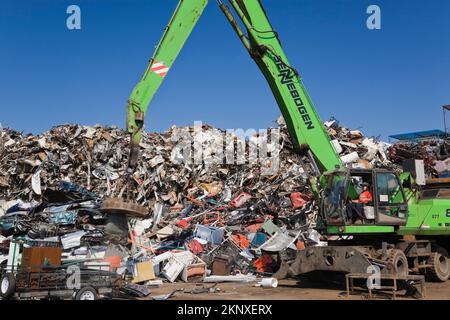 Kranlader mit Greiferarm und Magnet-Entladeanhänger vor dem Stapel von ausrangierten Autos, Haushaltsgeräten aus Metall und Industriegütern. Stockfoto