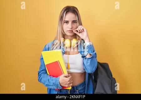 Junge blonde Frau, die einen Studentenrucksack trägt und Bücher mit Mund und Lippen wie Reißverschluss mit den Fingern hält. Geheim und still, Tabu-Reden Stockfoto