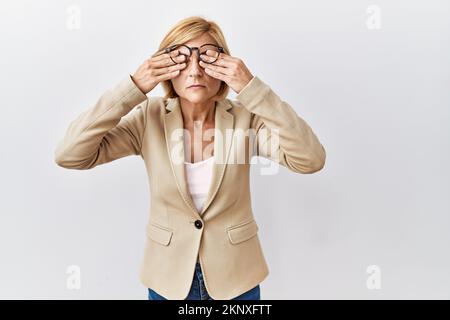 Blonde Geschäftsfrau mittleren Alters, die über einem isolierten Hintergrund steht und die Augen wegen Müdigkeit und Kopfschmerzen, Schläfrigkeit und Müdigkeit reibt. Sehstörungen Stockfoto
